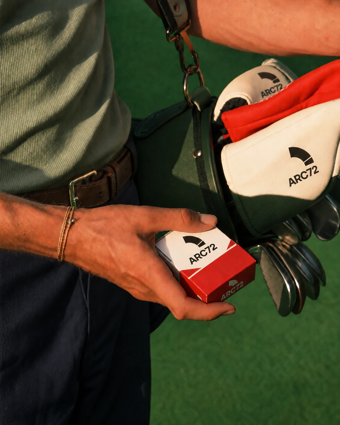Hånd som holder en ballpakke med ARC72-merket, ved siden av en golfbag med hodeovertrekk og jern.